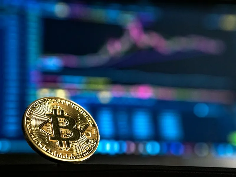 A physical Bitcoin coin in front of a market screen.